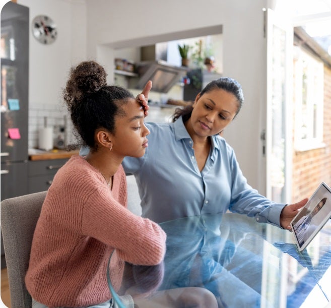 Mom feeling the forehead of her unwell teen with doctor on their tablet